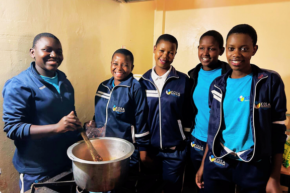 Five KGSA students in navy tracksuits smiling around a large cooking pot